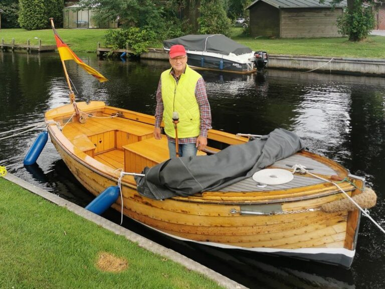 Älterer Mann mit roter Kappe steht auf einem Holzboot am Steg, hinten weht eine deutsche Flagge.