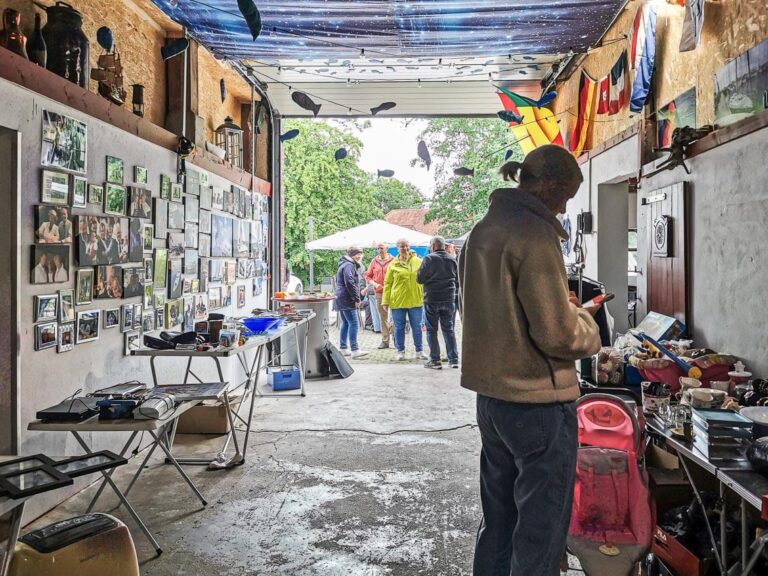 Menschen in einer offenen Garage mit Fotowand und Trödel, im Hintergrund Gespräche bei Pavillon im Freien.